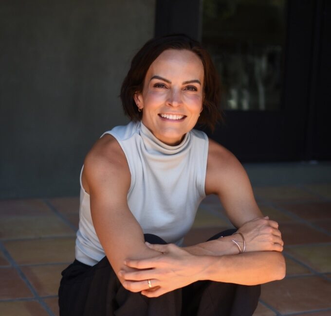 Trauma Solutions: A woman with short brown hair sits on a tiled floor, smiling, wearing a sleeveless white turtleneck and dark pants, with her arms resting on her knees.