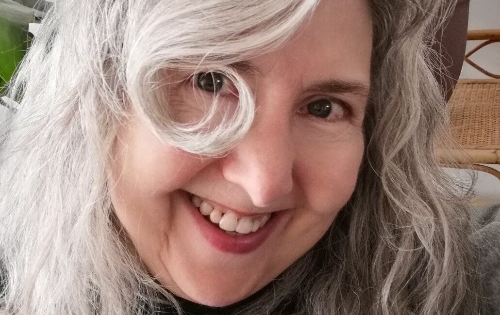 Trauma Solutions: A person with long, wavy gray hair smiles at the camera with plants visible in the background.