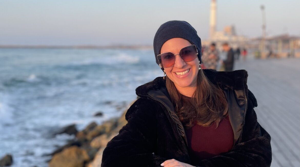 Trauma Solutions: Woman wearing sunglasses and a headscarf, smiling and leaning on a wooden railing by the seaside with a boardwalk and ocean in the background.