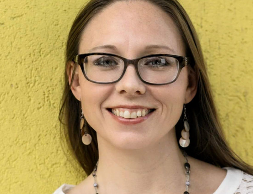A woman in glasses smiles in front of a yellow wall.