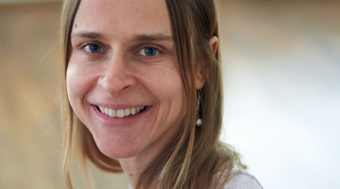Trauma Solutions: A woman with straight blonde hair and blue eyes, wearing a light-colored top, smiles at the camera in a softly lit indoor setting with a wooden floor.