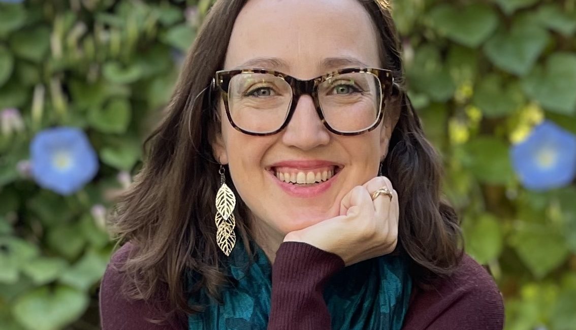 Sophie, a woman wearing glasses and a scarf, poses elegantly in front of flowers.