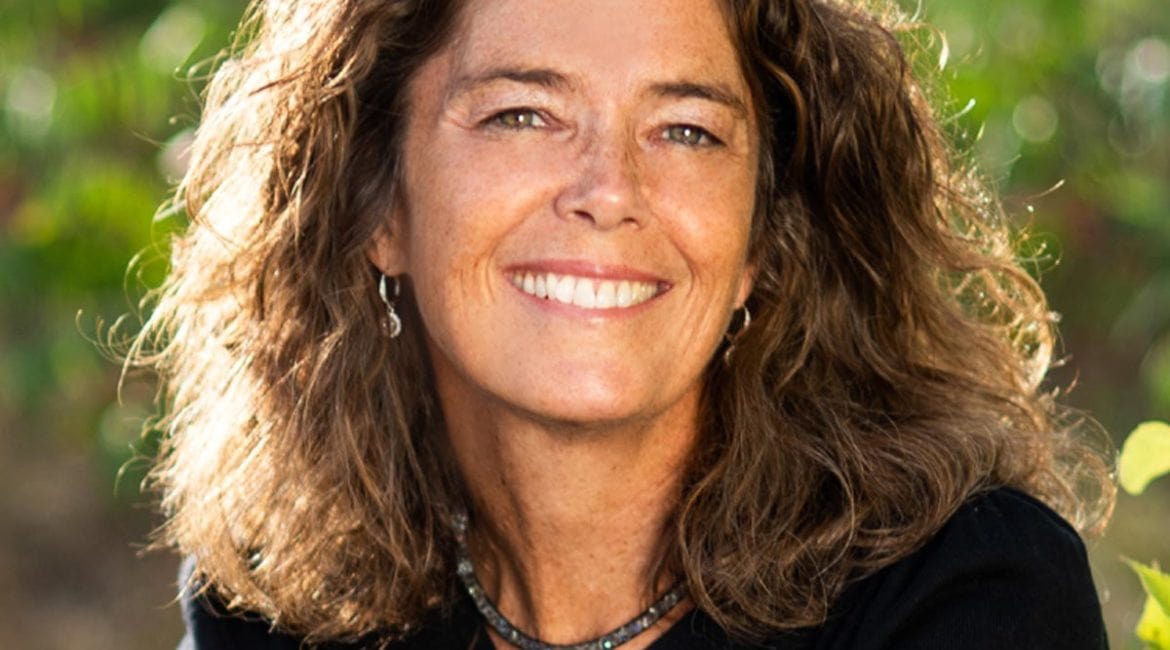 Liz George, a woman with curly hair, smiling in front of bushes.