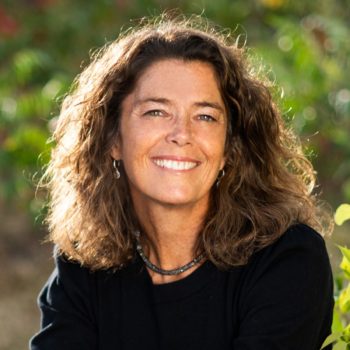 Liz George, a woman with curly hair, smiling in front of bushes.