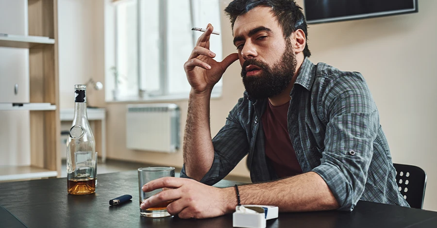 Trauma Solutions: A bearded man sitting at a table with a glass of liquor, a bottle, and a cigarette.