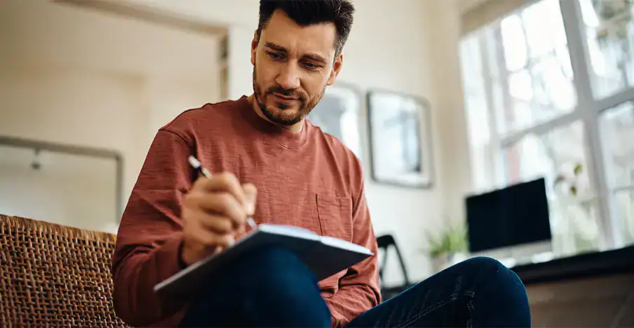 Trauma Solutions: Man wearing a red sweater sits indoors, writing in a notebook with a pen. A computer and window are visible in the background.