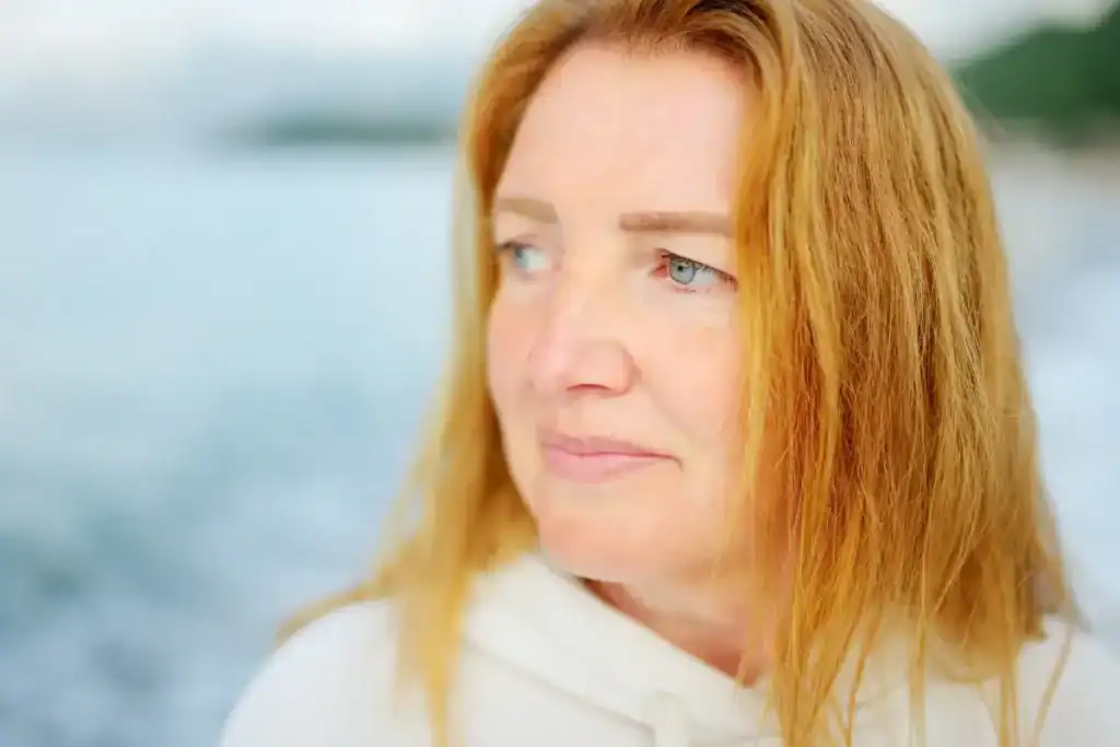 A woman stands at the shoreline with a melancholy expression, suggesting complex experiences of loss and somatic patterns of unresolved grief.