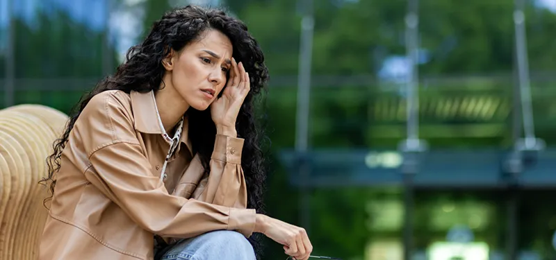 A woman sits outside with a distressed expression, resting her head in one hand. Her posture and facial tension suggest she is struggling with overwhelm or anxiety—reflecting the internal conflict often seen in trauma-related or encapsulated anxiety.