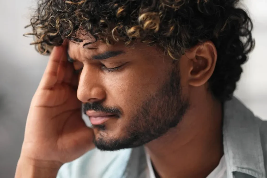 A young man with a pained expression holds his head, eyes closed in distress—illustrating the emotional and physiological toll of trauma-related anxiety and the unseen impact of encapsulated trauma on the nervous system.