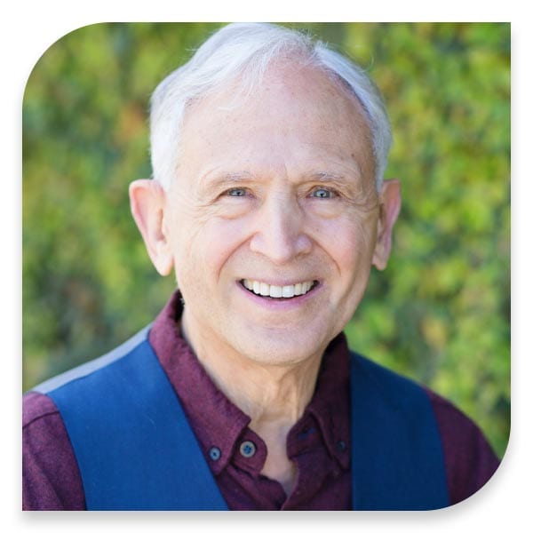 An elderly man smiling in front of his home's bushes.