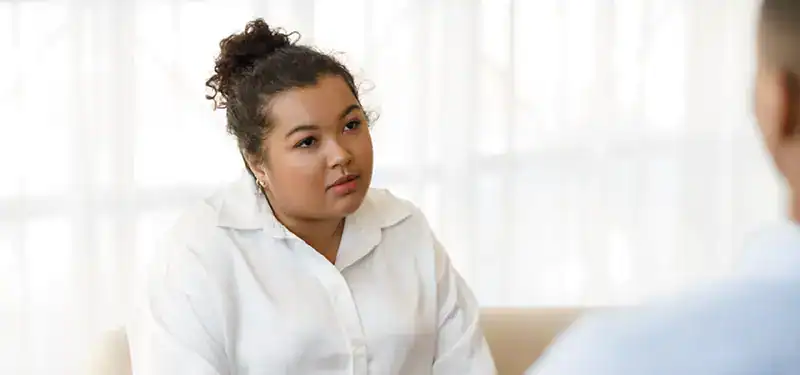 Trauma Solutions: A woman with curly hair, wearing a white shirt, sits and listens attentively with mindfulness to a person who is partially visible in the foreground.