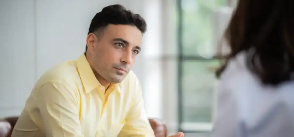 Trauma Solutions: A man in a yellow shirt sits indoors, practicing mindfulness as he attentively listens to a person in front of him whose back is to the camera.