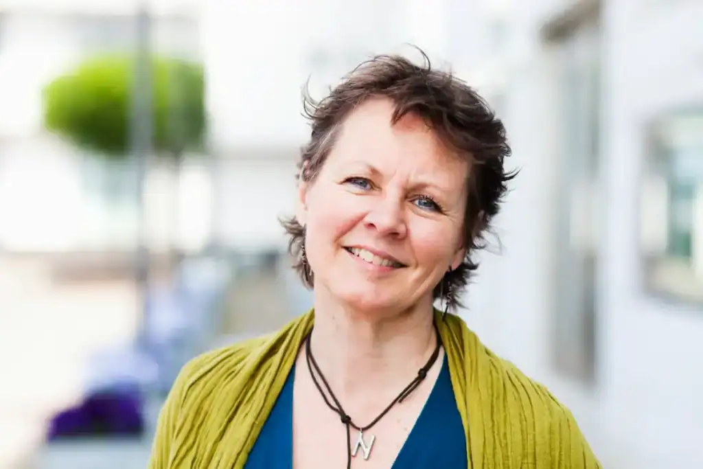 Trauma Solutions: A middle-aged woman with short brown hair smiles at the camera outdoors, radiating self-esteem while wearing a green scarf, blue top, and a necklace with a letter "N" pendant.