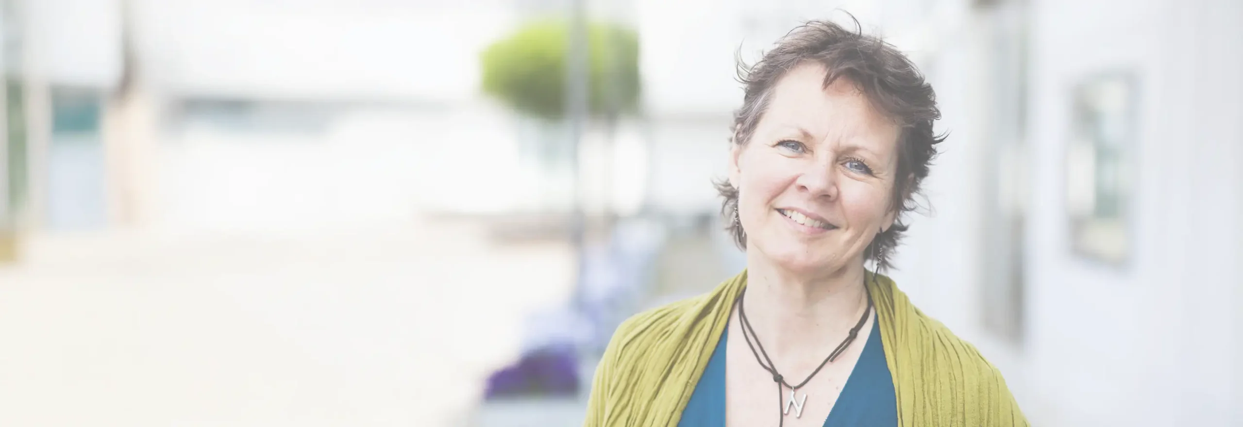 Trauma Solutions: A woman with short brown hair smiles at the camera, radiating self-esteem as she wears a green shawl and blue top, standing outdoors with a blurred background.