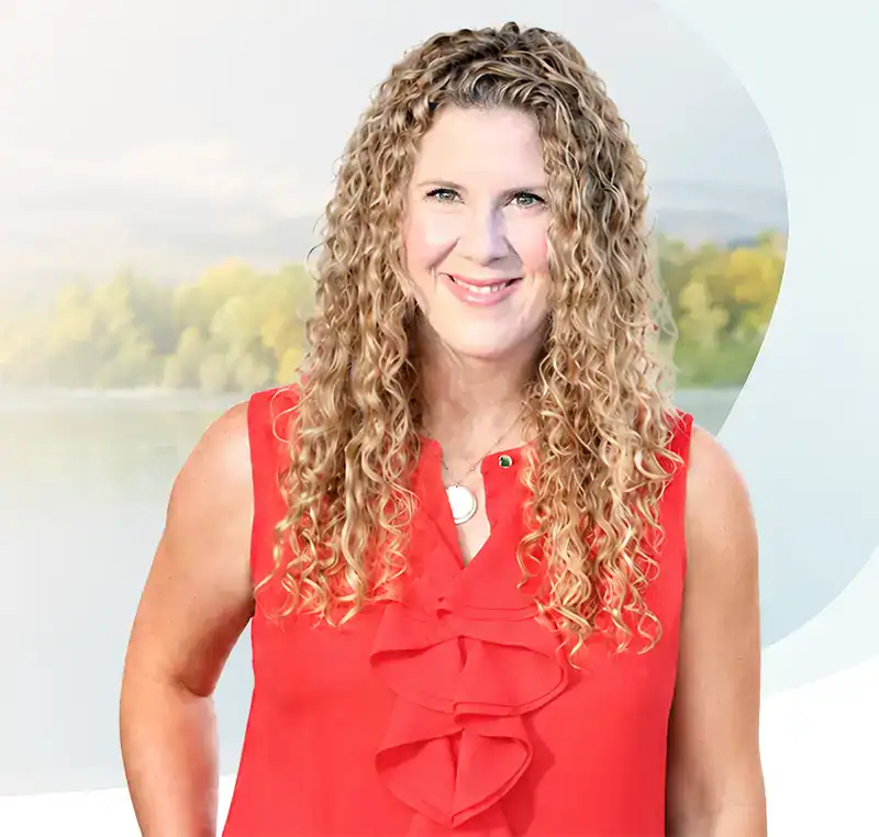 Trauma Solutions: Woman with long curly blonde hair wearing a sleeveless red blouse stands outdoors with a blurred background of trees and water.