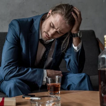 Trauma Solutions: A man in a suit sits on a couch, holding his head with one hand, with a glass of whiskey and an ashtray on the table in front of him.