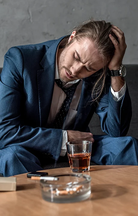 Trauma Solutions: A man in a suit sits slumped with his hand on his head, with a glass of whiskey and an ashtray on the table in front of him.