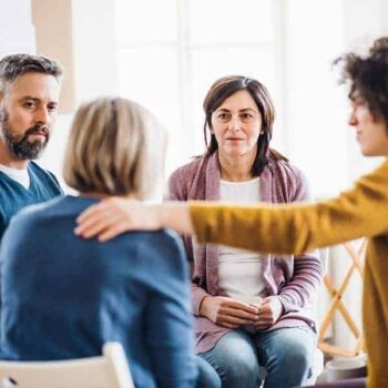 Trauma Solutions: Four adults sit in a circle having a group discussion; one person has a hand on another's shoulder, showing support.
