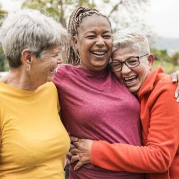 Trauma Solutions: Three older women stand outdoors, smiling and embracing each other closely, appearing happy and relaxed.
