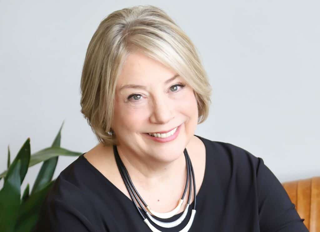 Trauma Solutions: A woman with short blonde hair, wearing a black top and a multi-strand necklace, smiles while sitting indoors with a plant in the background.