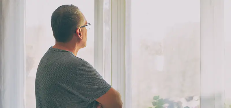 Trauma Solutions: A man wearing glasses stands indoors with arms crossed, looking out through large, bright windows, embodying a quiet mindfulness and self-esteem as he reflects on the world outside.