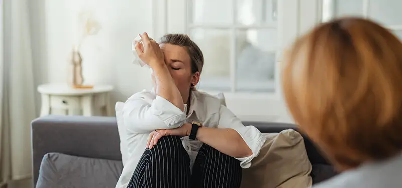Trauma Solutions: Person sitting on a couch wipes tears from their eyes while another person sits nearby, suggesting an emotional conversation or counseling setting focused on self-esteem and mindfulness.