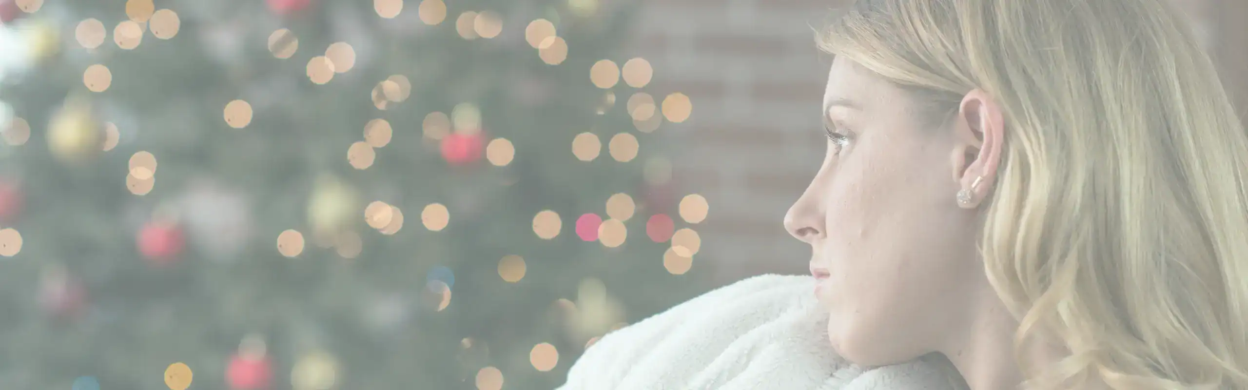 Trauma Solutions: Woman with blonde hair looks pensively to the side while sitting indoors; a decorated Christmas tree with lights and ornaments is blurred in the background, evoking a quiet moment of mindfulness and reflection on self-esteem during the festive season.