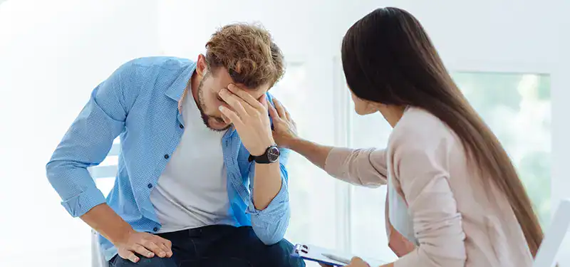 Trauma Solutions: A woman sits beside a man who appears distressed, with his head in his hand, and gently rests her hand on his shoulder in a supportive gesture.
