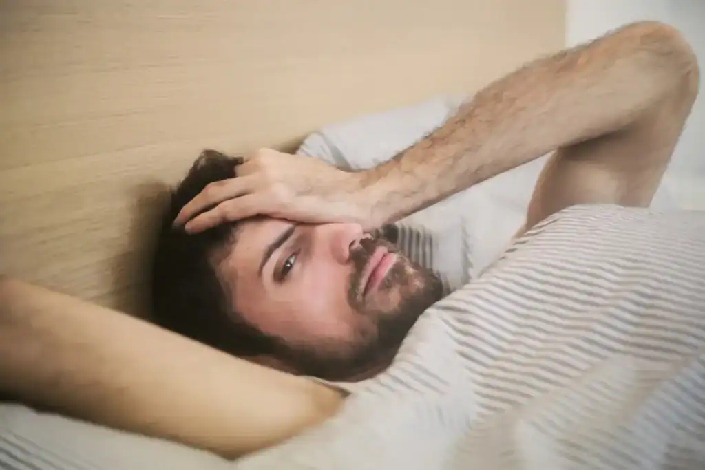 Trauma Solutions: A man lying in bed with one hand on his forehead, looking tired or pensive, as if reflecting on self-esteem or practicing mindfulness, partially covered by a striped blanket.