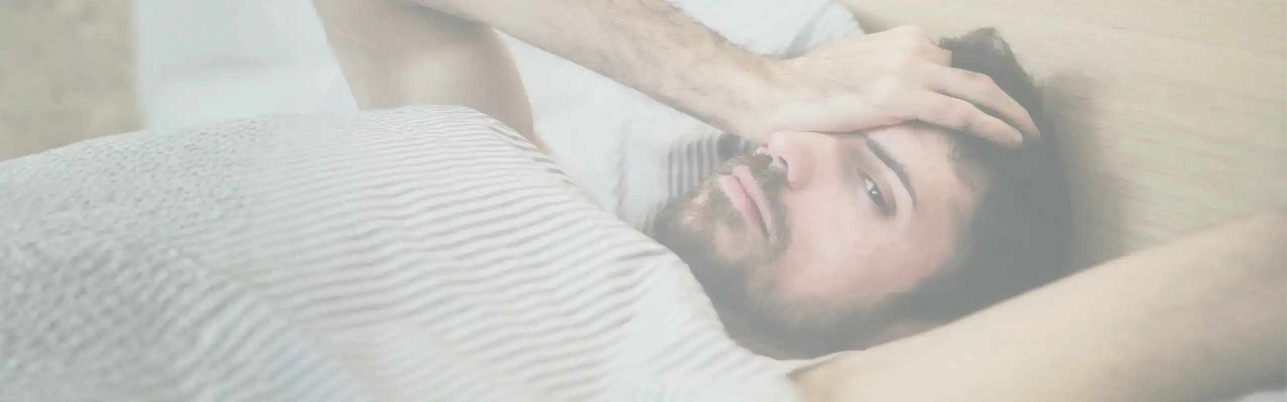 Trauma Solutions: A man lying in bed with a striped blanket, resting one hand on his forehead and looking slightly distressed or tired, reflecting on mindfulness and self-esteem. He lies awake, struggling to sleep. Often, hypervigilance in the nervous system prevents full relaxation and restoration.