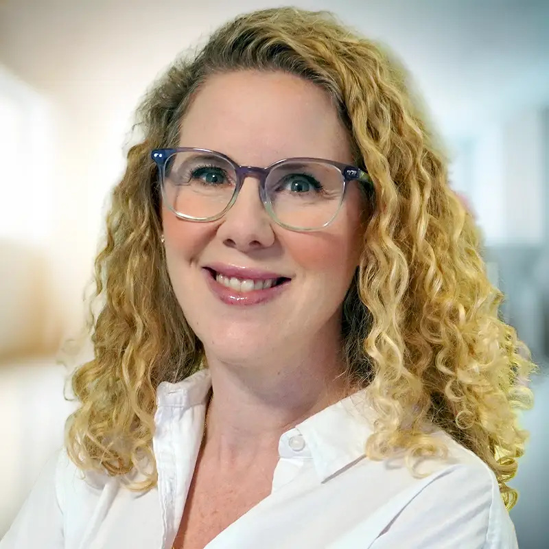 Trauma Solutions: Portrait of a smiling woman with curly blonde hair wearing glasses and a white collared shirt.
