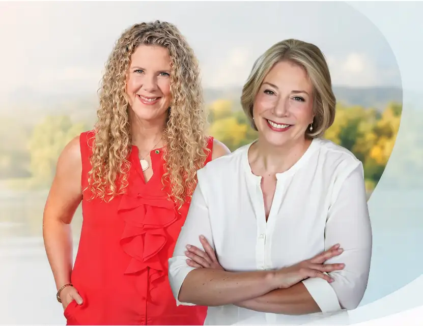 Trauma Solutions: Dr. Diane Poole Heller and Rose Viggiano are standing side by side, one in a sleeveless red blouse with curly hair, the other in a white blouse with short blonde hair, smiling in front of a blurred outdoor background.