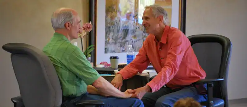 Trauma Solutions: Two older men sit facing each other in office chairs, holding hands and smiling, with a desk and framed artwork in the background.