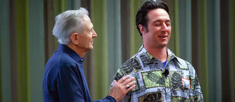 Trauma Solutions: An older man in a blue shirt places his hand on the shoulder of a younger man wearing a patterned shirt as they stand together indoors.
