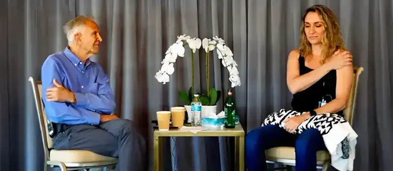 Trauma Solutions: A man and a woman sit facing each other on chairs with a small table between them holding drinks and flowers; both touch one shoulder, possibly demonstrating an exercise.