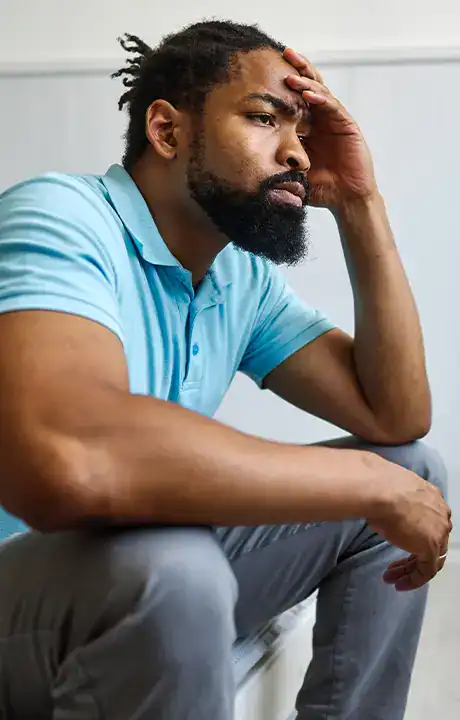 Trauma Solutions: A man with a beard sits on a bench wearing a light blue polo shirt and gray pants, resting his elbow on his knee and his hand on his forehead, looking thoughtful.