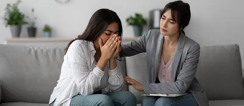 Trauma Solutions: Two women sit on a couch; one is visibly upset with her hands covering her face, while the other offers comfort with a hand on her shoulder.
