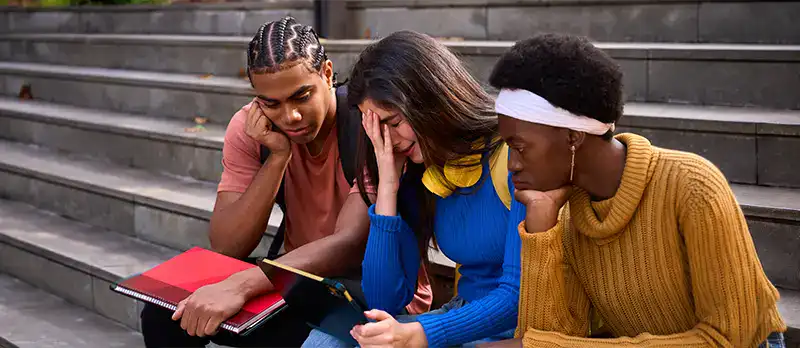 Trauma Solutions: Three students sitting on outdoor steps look at a tablet with concerned expressions, holding notebooks and resting their heads on their hands.