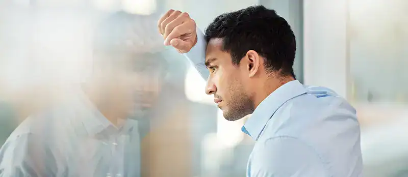 Trauma Solutions: A man in a light blue shirt leans his forehead and arm against a glass window, looking thoughtful or concerned.